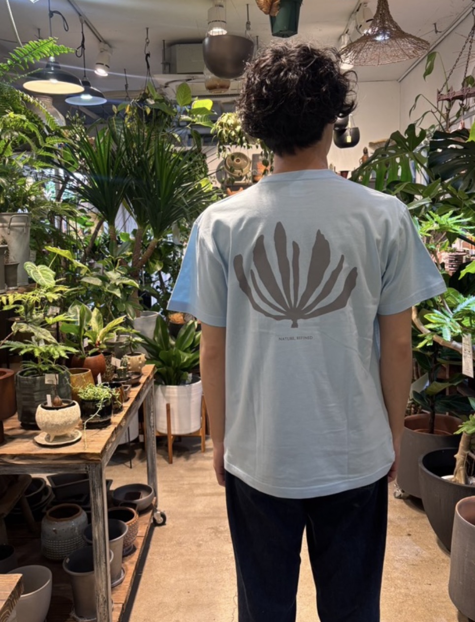 植物に囲まれた店舗でGXアートTシャツ（ライトブルー）の背面を着用したスタイリング